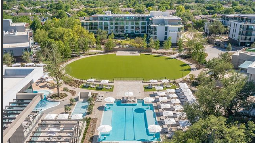Aerial view of The Village - Dallas TX. Swiming pool with white umbrellas around, near a large circle of green grass. Trees and buildings around.