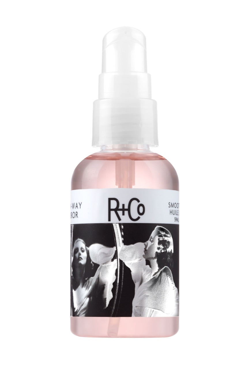 A spray bottle of R+Co Two-Way Mirror Smoothing Oil with a label featuring a black and white photograph of a person looking into a mirror