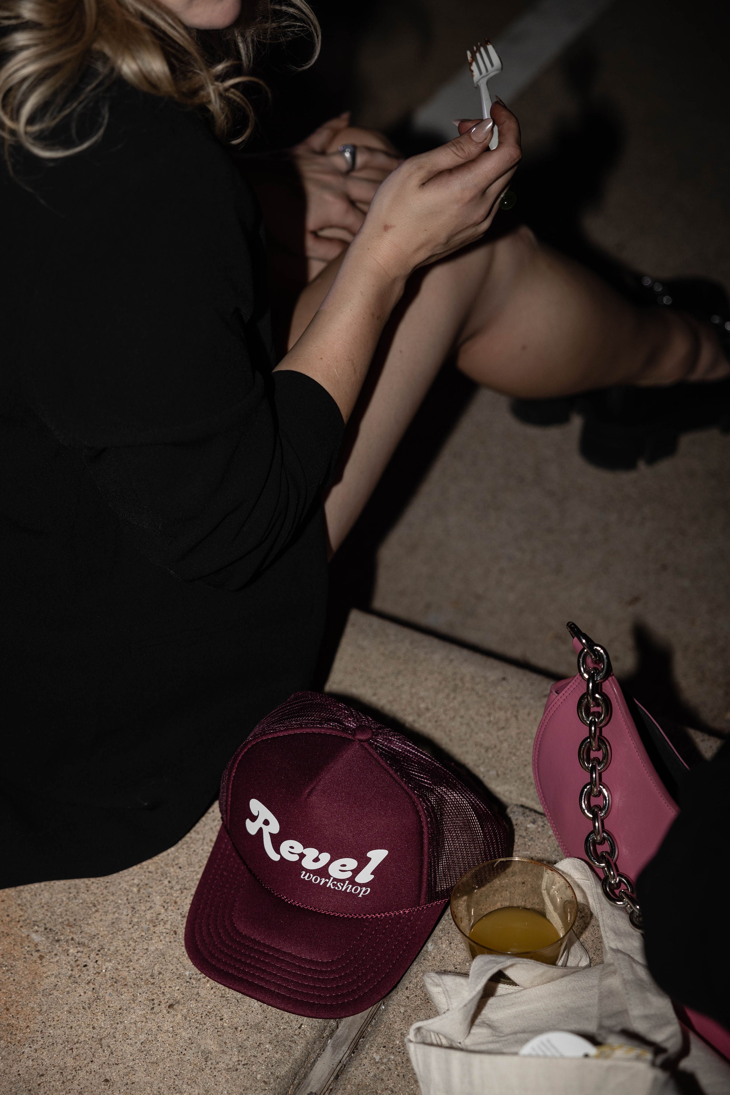 A lady sitting on the sidewalk during Revel's grand opening party with maroon Revel Workshop trucker hat prominently displayed