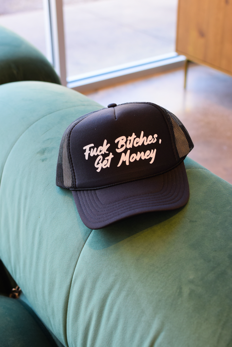 This image features a bold and attention-grabbing trucker hat with the phrase "Fuck Bitches, Get Money" prominently displayed. The hat, with a black brim and crown, offers a casual and rugged look, suitable for those who want to make a statement with their fashion choices.