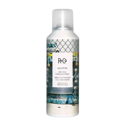 A white spray bottle of R+Co HALFPIPE  dry wax finishing spray with a label showing skateboards and chain link fence