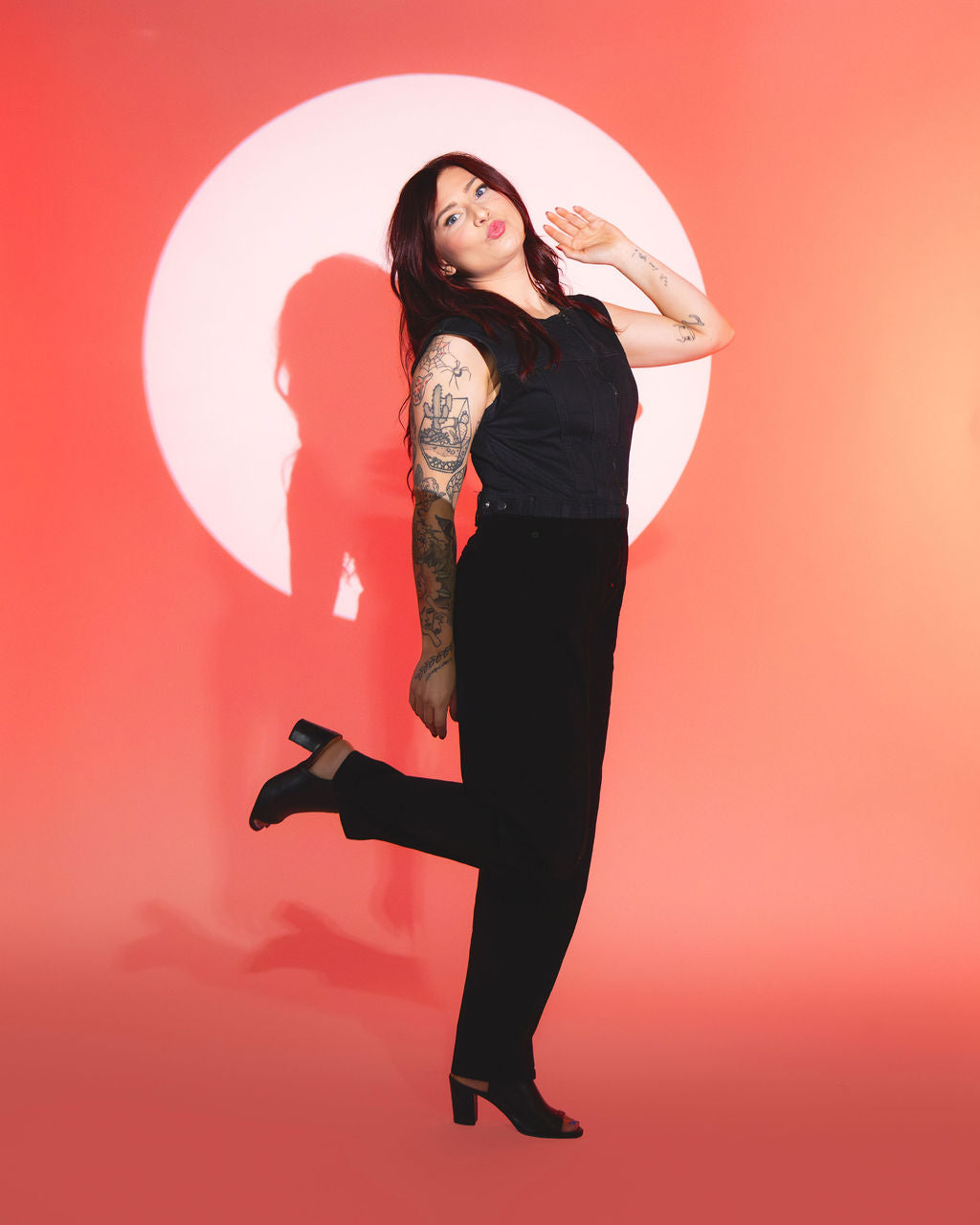 Kaylee playfully standing in spotlight wearing black outfit against orange background