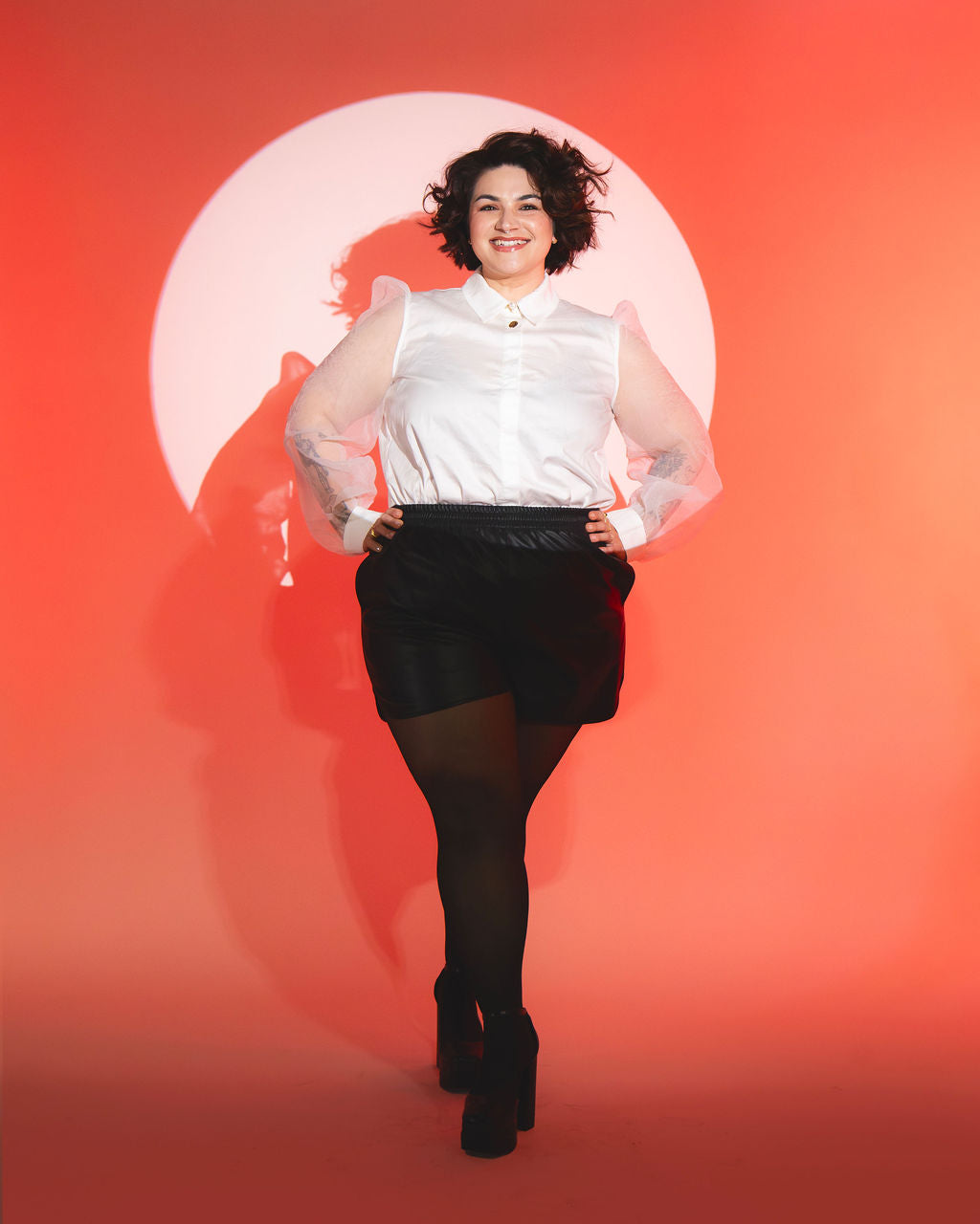 Meagan standing in spotlight wearing white blouse against orange background