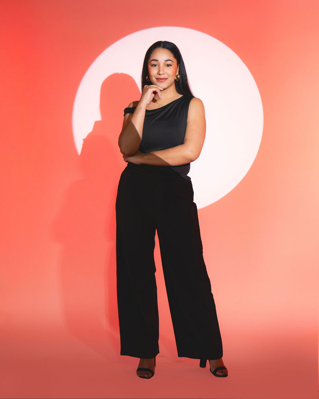 Trinity standing in spotlight wearing black outfit against orange background