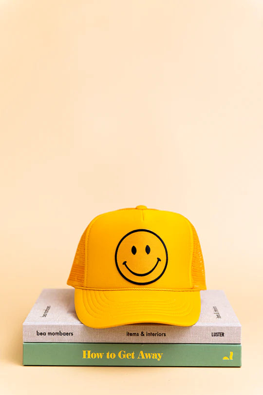 Yellow trucker hat with smiley face design on a stack of books against a beige background
