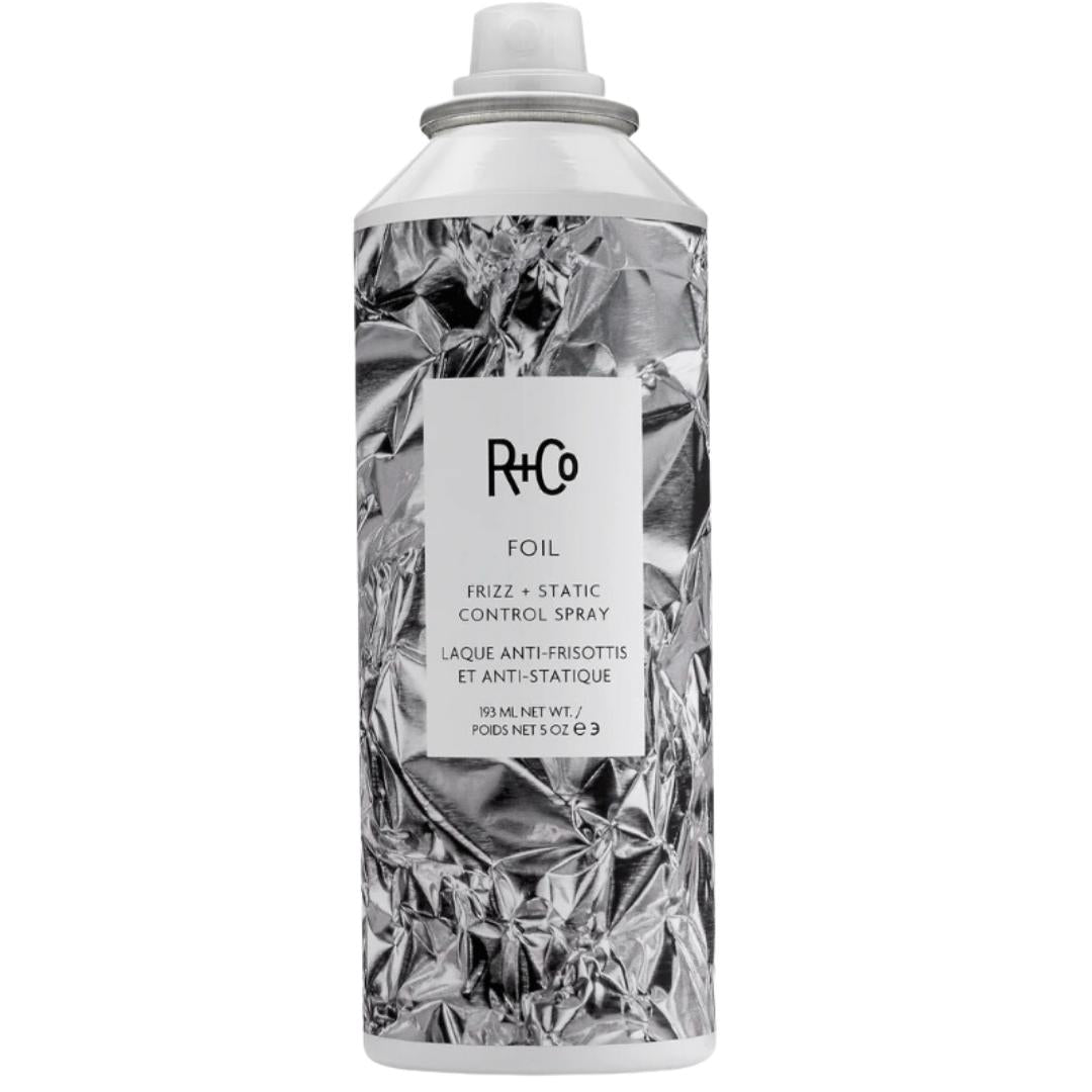 A bottle of R+Co Foil Frizz + Static Control Spray with a silver and white label, designed for hair styling.