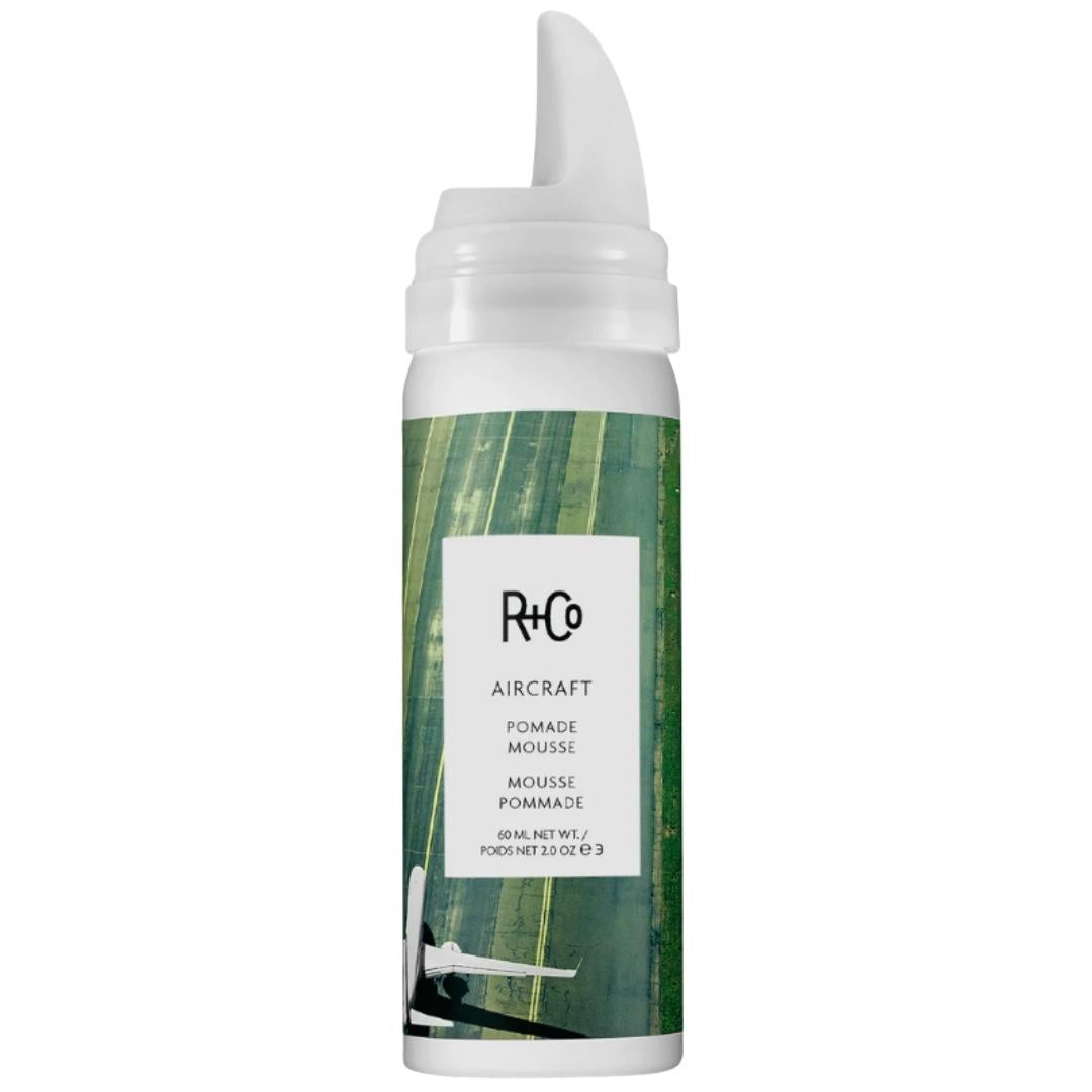 A bottle of AIRCRAFT Pomade Mousse by R+Co with a green label and runway imagery.