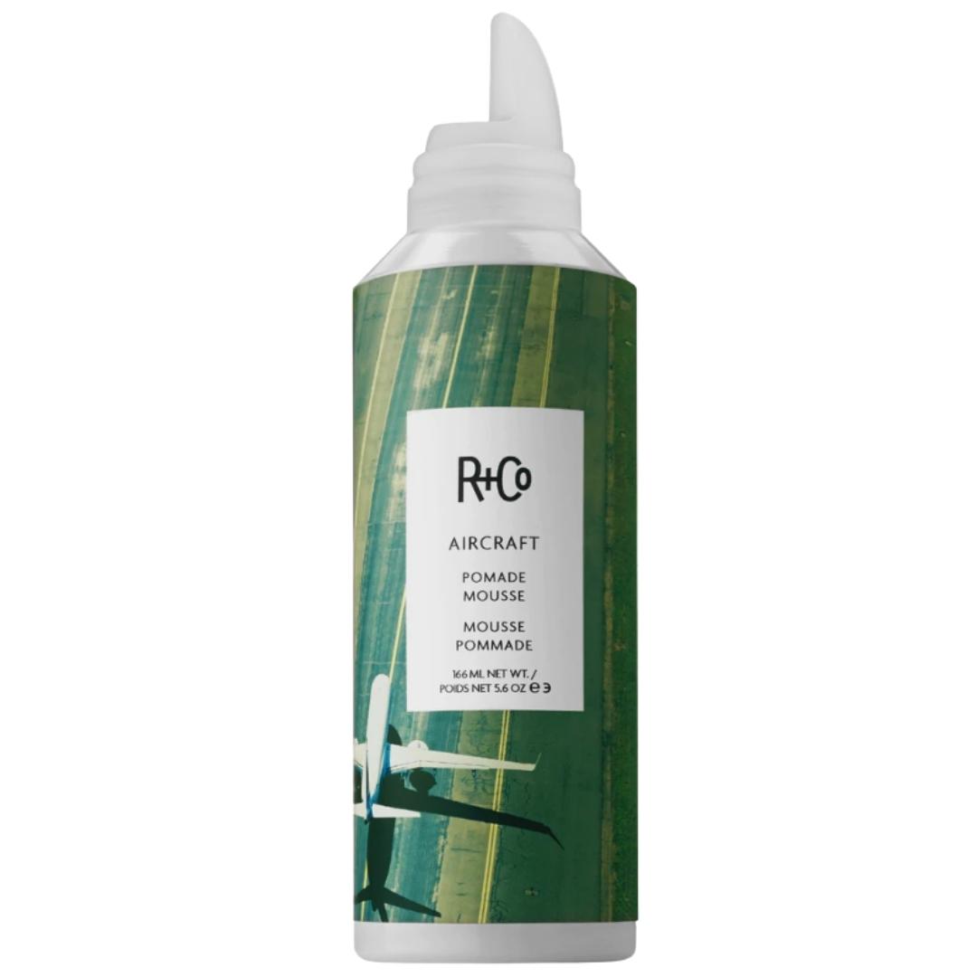 A bottle of AIRCRAFT Pomade Mousse by R+Co with a green label and runway imagery.